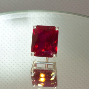 Puede incluir: Una piedra preciosa de color rojo intenso engastada en un anillo de plata. El anillo está en un soporte de exhibición.