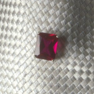May include: A square-shaped red gemstone with a gold setting. The gemstone is on a white textured background.