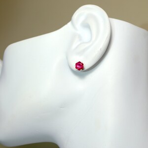 May include: A pair of small, pink stud earrings with a square shape. The earrings are shown on a white mannequin head.