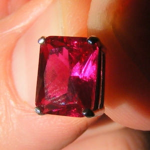 May include: A deep red gemstone set in a silver bezel setting. The gemstone is a rectangular emerald cut.