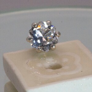 May include: A clear, faceted, round gemstone with a silver setting. The gemstone is on a white, ceramic display stand.