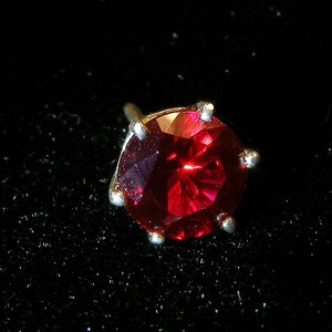 Op de afbeelding: Enkele oorbel met een grote, ronde, gefacetteerde, dieprode edelsteen. De edelsteen is gezet in een zilverkleurig metaal met zes klauwen. De oorbel is geplaatst tegen een zwarte achtergrond.