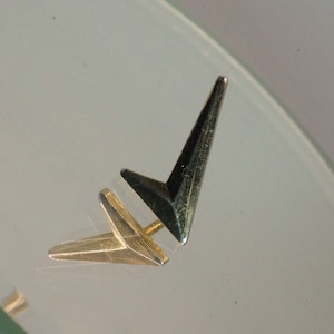 Puede incluir: Un par de pendientes de flecha de oro y negro. Los pendientes están hechos de metal y tienen un diseño simple y moderno.