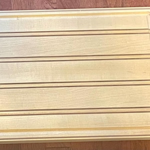 May include: Rectangular wooden cutting board with a light wood grain. The board has a recessed border and horizontal grooves. The wood has a natural, light color with darker brown accent lines.