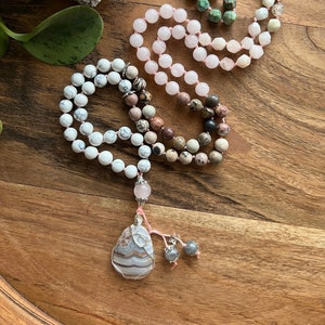 May include: Three gemstone mala bead necklaces in shades of white, pink, and green, with a wire-wrapped pendant. The necklaces are displayed on a wooden surface, showcasing the natural colors and textures of the beads and pendant.