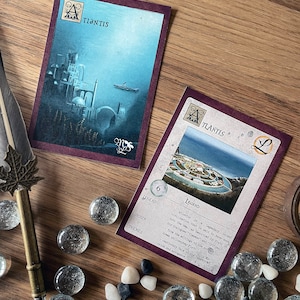 K&ouml;nnte beinhalten: Zwei Spielkarten mit einem violetten Hintergrund und goldenen Akzenten. Die Karten zeigen eine Illustration einer Stadt und den Text "TLANTIS" und "ATLANTIS". Die Karte rechts enth&auml;lt eine Legende &uuml;ber die Stadt Atlantis.