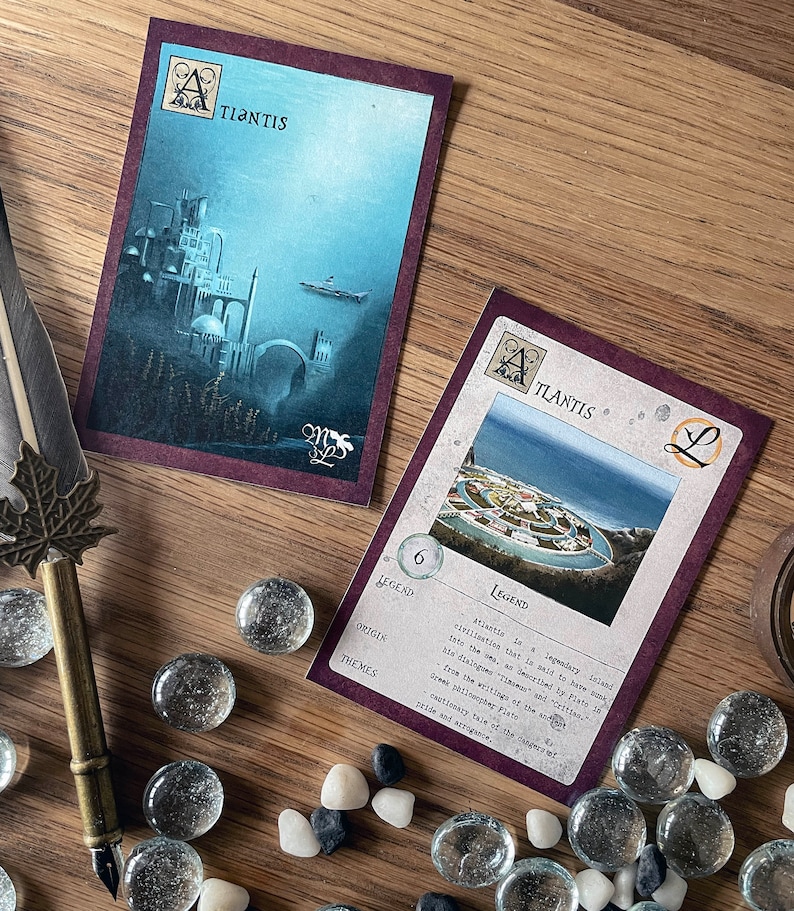 K&ouml;nnte beinhalten: Zwei Spielkarten mit einem violetten Hintergrund und goldenen Akzenten. Die Karten zeigen eine Illustration einer Stadt und den Text "TLANTIS" und "ATLANTIS". Die Karte rechts enth&auml;lt eine Legende &uuml;ber die Stadt Atlantis.