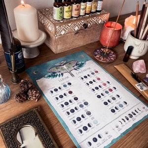 Pu&ograve; includere: Un calendario lunare blu e bianco con un'illustrazione di falena. Il calendario mostra le fasi lunari per ogni mese dell'anno. Il calendario &egrave; su un tavolo di legno con vari oggetti, tra cui candele, oli essenziali e cristalli.
