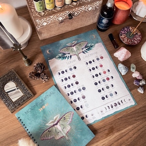 Pu&ograve; includere: Un diario lunare blu-verde con un designo di falena lunare in foglia d'oro e le parole "Moon Journal" sulla copertina. Il diario &egrave; aperto a una pagina con un calendario delle fasi lunari dell'anno. Il calendario &egrave; stampato su uno sfondo blu-verde con un designo di falena lunare in foglia d'oro.