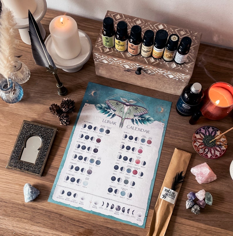 Pu&ograve; includere: Un calendario lunare con un'illustrazione ad acquerello di una falena su uno sfondo blu-verde. Il calendario mostra le fasi lunari per ogni mese dell'anno. Il calendario &egrave; circondato da vari oggetti, tra cui candele, oli essenziali, cristalli e incenso.