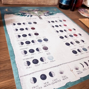 Pu&ograve; includere: Un calendario lunare color verde acqua e bianco con un'illustrazione di una falena. Il calendario mostra le fasi lunari per ogni mese dell'anno. Il testo sul calendario include i nomi dei mesi, le date delle fasi lunari e i nomi delle lune piene.