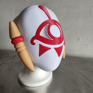 Puede incluir: Una m&aacute;scara blanca con detalles rojos, que presenta un dise&ntilde;o de ojo estilizado y formas triangulares. La m&aacute;scara tiene piezas laterales de color beige con detalles rojos. Se muestra sobre un soporte blanco, ideal para cosplay o disfraces.