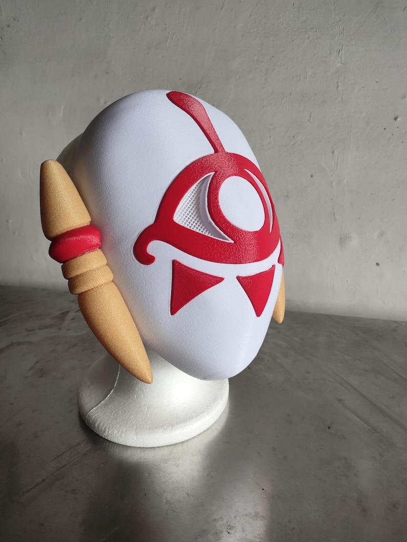 Puede incluir: Una m&aacute;scara blanca con detalles rojos, que presenta un dise&ntilde;o de ojo estilizado y formas triangulares. La m&aacute;scara tiene piezas laterales de color beige con detalles rojos. Se muestra sobre un soporte blanco, ideal para cosplay o disfraces.