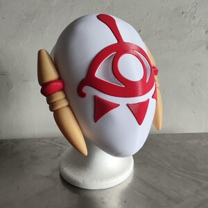 Puede incluir: Una m&aacute;scara blanca con detalles rojos, que presenta un dise&ntilde;o de ojo y formas triangulares. La m&aacute;scara tiene piezas laterales de color beige y se muestra sobre un soporte blanco. Probablemente para cosplay o disfraces.