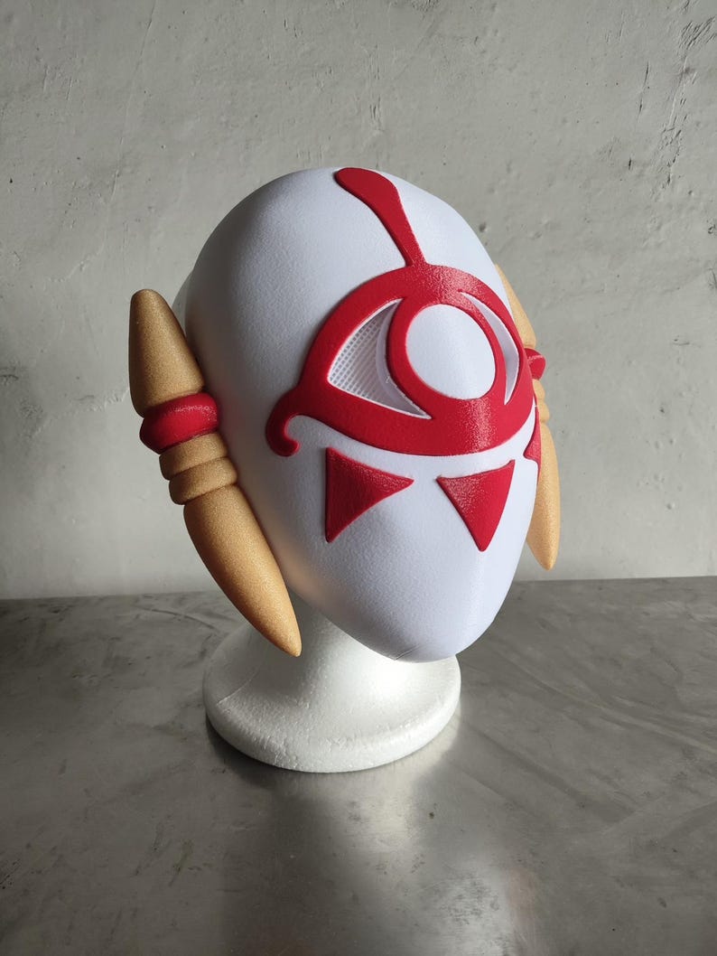 Puede incluir: Una m&aacute;scara blanca con detalles rojos, que presenta un dise&ntilde;o de ojo y formas triangulares. La m&aacute;scara tiene piezas laterales de color beige y se muestra sobre un soporte blanco. Probablemente para cosplay o disfraces.