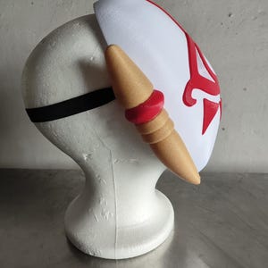 Puede incluir: Una m&aacute;scara blanca con detalles rojos y un elemento decorativo lateral. La m&aacute;scara tiene forma curva y se asegura con una banda el&aacute;stica negra. El elemento lateral es de color beige con detalles rojos.