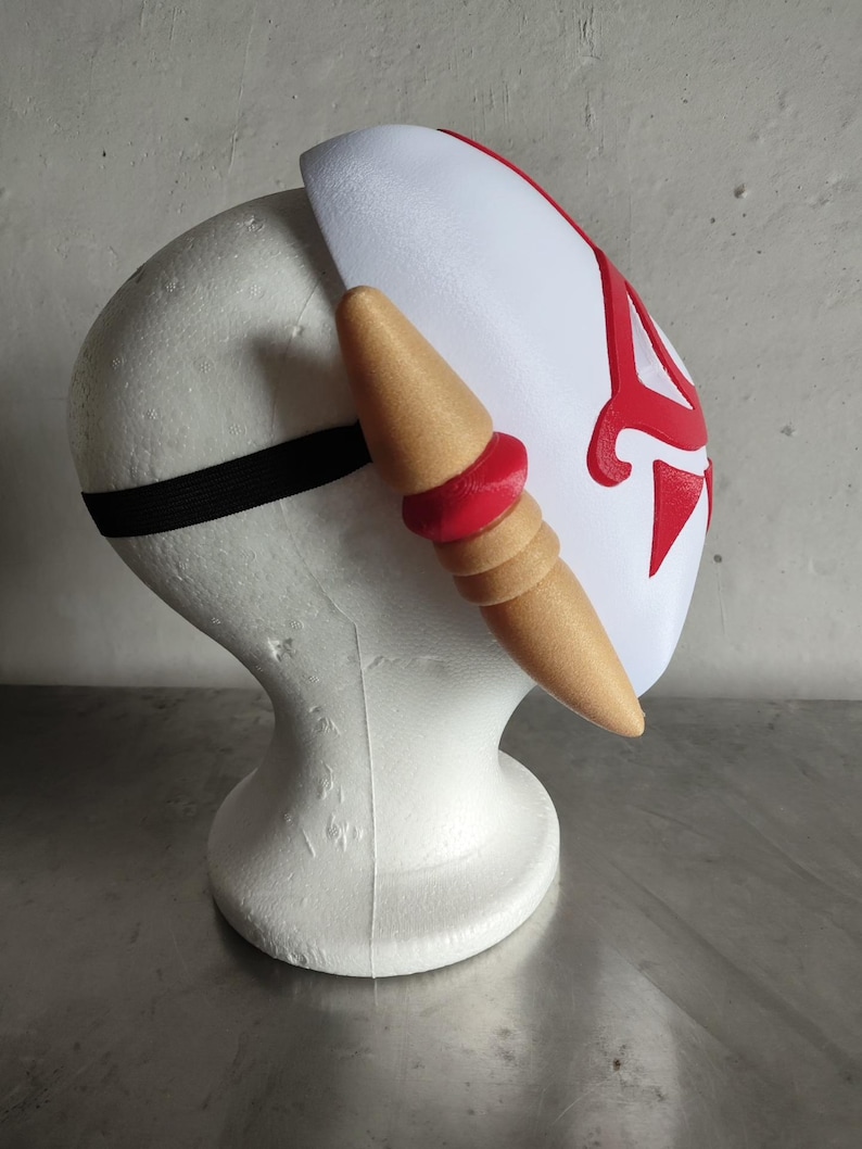 Puede incluir: Una m&aacute;scara blanca con detalles rojos y un elemento decorativo lateral. La m&aacute;scara tiene forma curva y se asegura con una banda el&aacute;stica negra. El elemento lateral es de color beige con detalles rojos.