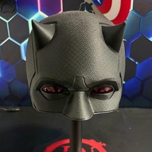 Peut inclure: Un masque de super-héros noir avec des détails texturés et des cornes pointues. Le masque présente des lentilles rouges et est exposé sur un support noir. Le masque est une réplique d'un personnage de bande dessinée.