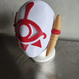 Puede incluir: Una m&aacute;scara blanca con un dise&ntilde;o de ojo rojo y detalles rojos. La m&aacute;scara tiene dos piezas laterales de color beige con bandas rojas. La m&aacute;scara est&aacute; sobre un soporte blanco.