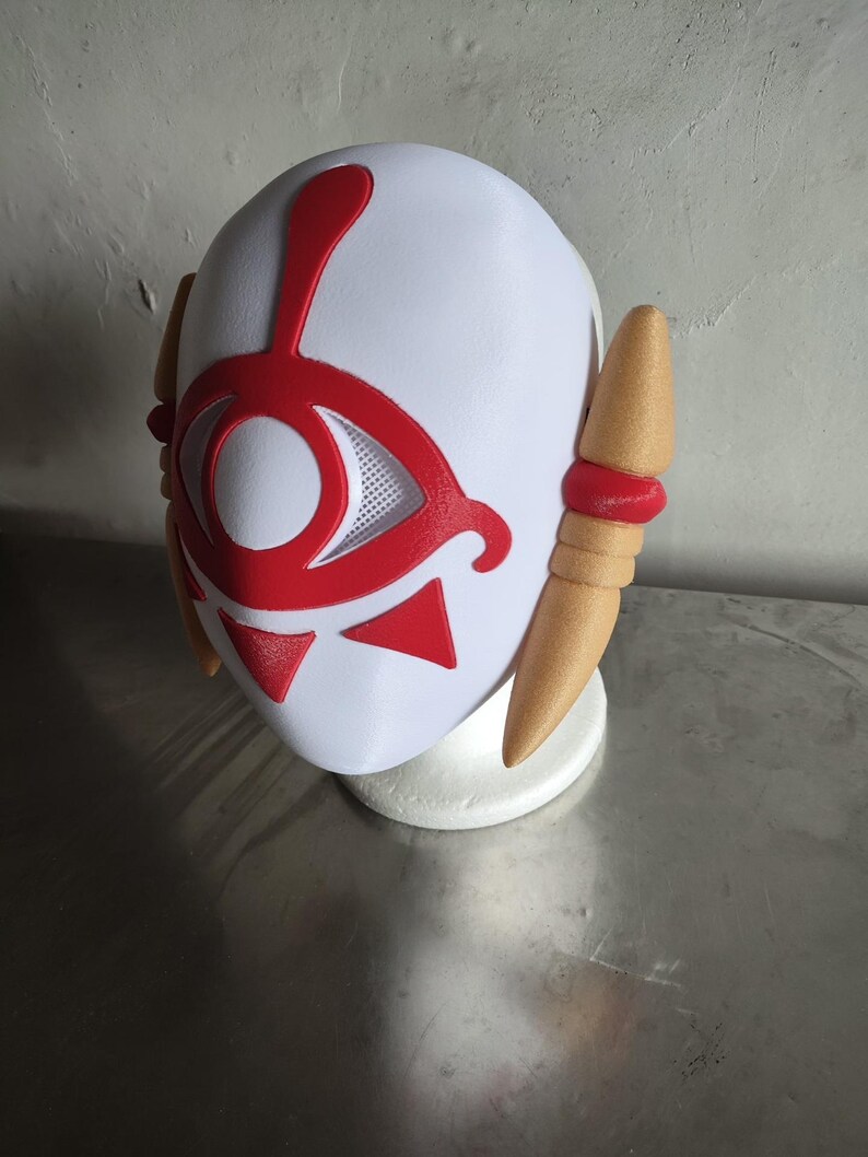 Puede incluir: Una m&aacute;scara blanca con un dise&ntilde;o de ojo rojo y detalles rojos. La m&aacute;scara tiene dos piezas laterales de color beige con bandas rojas. La m&aacute;scara est&aacute; sobre un soporte blanco.