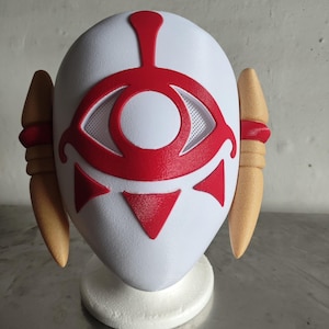 Puede incluir: Una m&aacute;scara blanca con un dise&ntilde;o de ojo rojo y detalles. La m&aacute;scara presenta un s&iacute;mbolo de ojo central con un &aacute;rea de ojo de malla, tri&aacute;ngulos rojos y una l&iacute;nea vertical roja. Dos formas en forma de cuerno, de color beige, est&aacute;n en los lados, con bandas rojas.