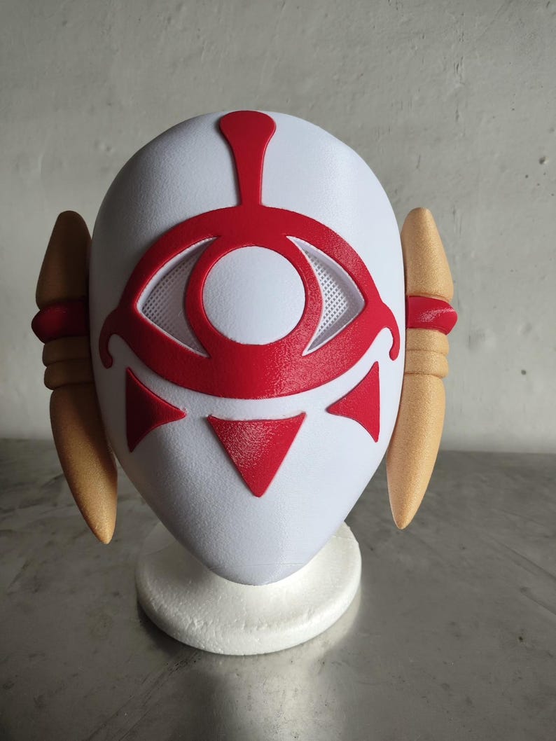 Puede incluir: Una m&aacute;scara blanca con un dise&ntilde;o de ojo rojo y detalles. La m&aacute;scara presenta un s&iacute;mbolo de ojo central con un &aacute;rea de ojo de malla, tri&aacute;ngulos rojos y una l&iacute;nea vertical roja. Dos formas en forma de cuerno, de color beige, est&aacute;n en los lados, con bandas rojas.