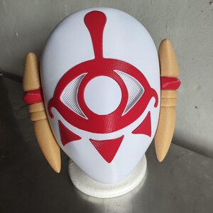 Puede incluir: Una m&aacute;scara blanca con un dise&ntilde;o de ojo rojo y detalles, con dos piezas laterales de color beige con bandas rojas. La m&aacute;scara es probablemente para cosplay o disfraces.
