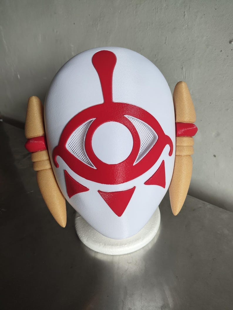 Puede incluir: Una m&aacute;scara blanca con un dise&ntilde;o de ojo rojo y detalles, con dos piezas laterales de color beige con bandas rojas. La m&aacute;scara es probablemente para cosplay o disfraces.