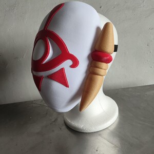 Puede incluir: Una m&aacute;scara blanca con detalles rojos, que presenta un dise&ntilde;o estilizado. La m&aacute;scara tiene una forma curvada y est&aacute; colocada sobre un soporte blanco. Un elemento en forma de cuerno, de color beige, se extiende desde el lateral, con una banda roja. Una correa negra es visible.