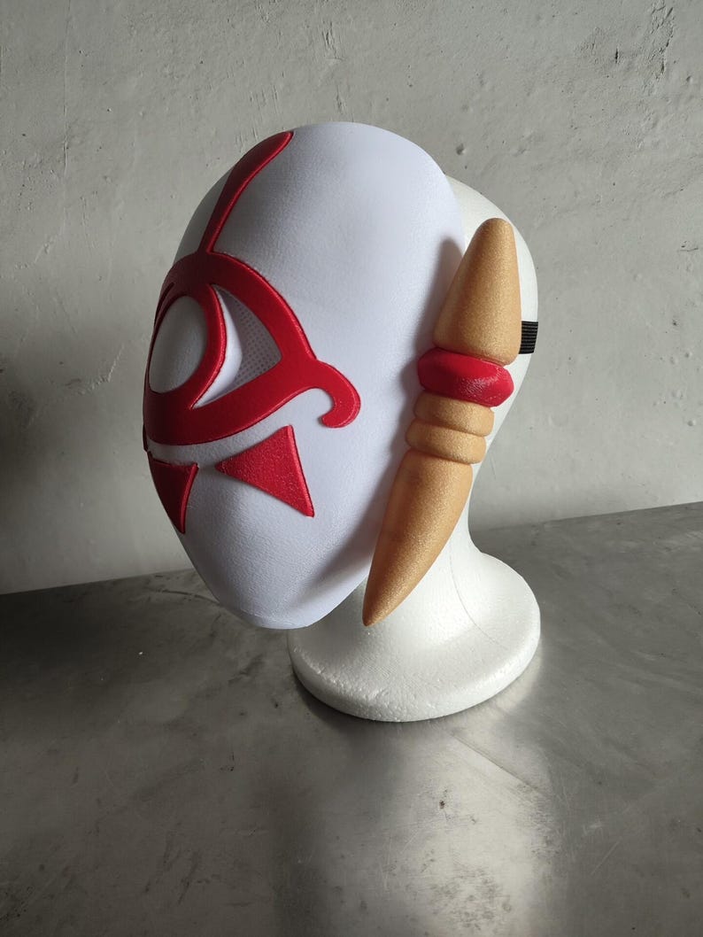 Puede incluir: Una m&aacute;scara blanca con detalles rojos, que presenta un dise&ntilde;o estilizado. La m&aacute;scara tiene una forma curvada y est&aacute; colocada sobre un soporte blanco. Un elemento en forma de cuerno, de color beige, se extiende desde el lateral, con una banda roja. Una correa negra es visible.