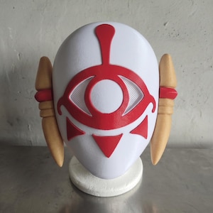 Puede incluir: Una m&aacute;scara blanca con un dise&ntilde;o de ojo rojo y detalles en rojo. La m&aacute;scara tiene dos piezas laterales de color beige y est&aacute; montada sobre una base blanca. El dise&ntilde;o del ojo presenta un centro de malla y formas triangulares debajo.