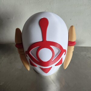 Puede incluir: Una m&aacute;scara blanca con un dise&ntilde;o de ojo rojo y detalles rojos. La m&aacute;scara tiene dos protuberancias en forma de oreja de color beige a cada lado. El dise&ntilde;o del ojo est&aacute; centrado y tiene una textura similar a una malla.
