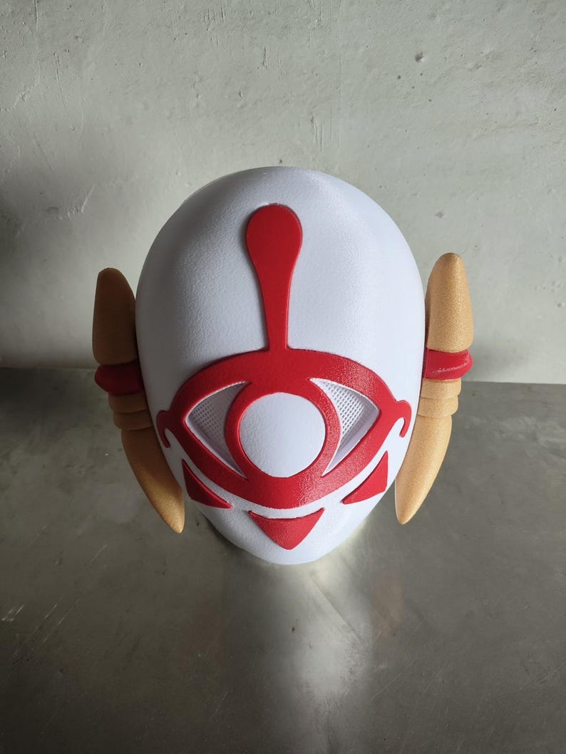 Puede incluir: Una m&aacute;scara blanca con un dise&ntilde;o de ojo rojo y detalles rojos. La m&aacute;scara tiene dos protuberancias en forma de oreja de color beige a cada lado. El dise&ntilde;o del ojo est&aacute; centrado y tiene una textura similar a una malla.
