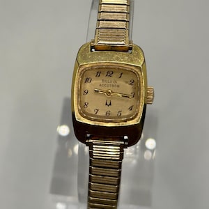 May include: A gold-tone Bulova Accutron wristwatch featuring a rectangular case with rounded edges, a light-colored dial with black Arabic numerals, and a flexible metal expansion band. The watch face displays the text Bulova Accutron.