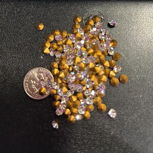 May include: A collection of gold-coloured, pyramid-shaped studs and iridescent rhinestones. A U.S. quarter coin is included for size reference. The studs and rhinestones are scattered on a black surface, creating a sparkling effect.