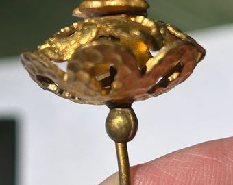 May include: A gold-toned hat pin with a decorative, openwork design. The pin features a small, rounded top and a detailed, openwork cap. The pin's stem is slender and ends in a small, spherical detail.