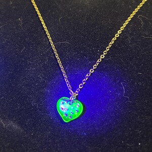 May include: A gold-coloured chain necklace with a heart-shaped pendant. The pendant is a vibrant green colour with small, sparkling accents. The necklace is displayed against a dark blue background.