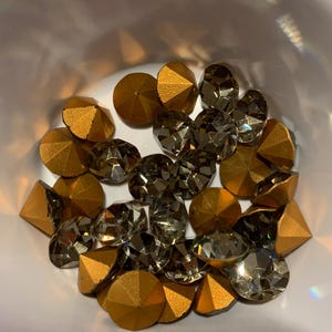 May include: A collection of gold and clear rhinestones in a bowl. The rhinestones are faceted and have a pointed top.