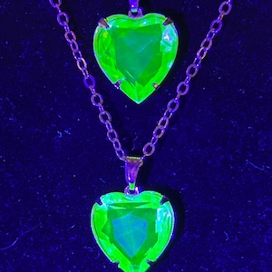 May include: Two necklaces with heart-shaped pendants. The pendants are a vibrant neon green, set in a gold-colored metal. The chains are delicate and also gold-toned, against a dark blue background.