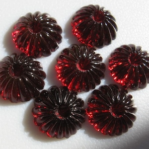 May include: Seven dark red glass cabochons with a fluted design. The cabochons are arranged in a circle on a white surface.