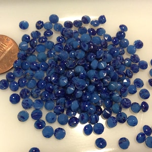May include: A large pile of small, round, blue glass beads with gold flecks. The beads are scattered on a white surface.
