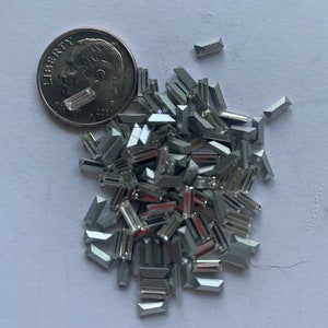 May include: A pile of silver-toned, rectangular-shaped rhinestones. The rhinestones are scattered on a white surface. A US dime coin is included for scale.