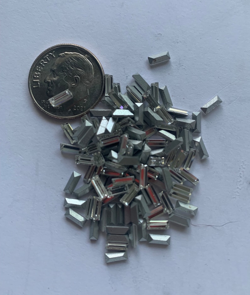 May include: A pile of silver-toned, rectangular-shaped rhinestones. The rhinestones are scattered on a white surface. A US dime coin is included for scale.