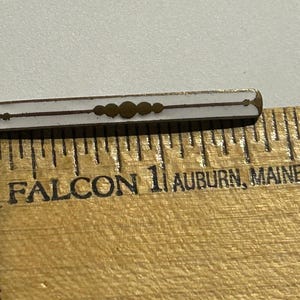 May include: A small, rectangular pin with a white enamel center and gold-colored accents. The pin is approximately 1 inch long and is placed next to a wooden ruler. The ruler is marked with inch increments and the text "FALCON 1 AUBURN, MAINE 2".