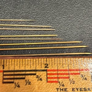 May include: A set of ten gold-colored metal pins with pointed ends and a small loop at the top. The pins are arranged in order of size, with the smallest pin at the top and the largest pin at the bottom. The pins are measured against a ruler, with the largest pin measuring approximately 2 inches in length.