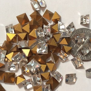 May include: A pile of gold and clear glass pyramid-shaped rhinestones. The rhinestones are scattered on a white surface. A US dime is included for scale.