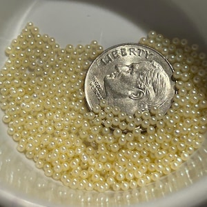 May include: A close-up shot of a white bowl filled with small, cream-colored beads. A U.S. dime is partially visible, resting among the beads. The dime features the profile of a person and the word "LIBERTY".