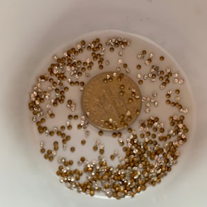 May include: A white bowl filled with small, gold and clear rhinestones. A coin is in the center of the bowl, surrounded by the rhinestones. The bowl is set against a white background. The rhinestones are likely used for crafts.