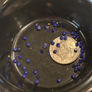 May include: A small black bowl filled with blue and gold glass beads. A US dime coin is in the center of the bowl.