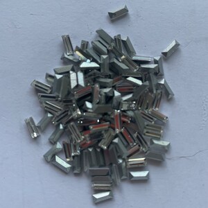 May include: A pile of silver-colored, rectangular-shaped rhinestones. The rhinestones are scattered on a white surface.
